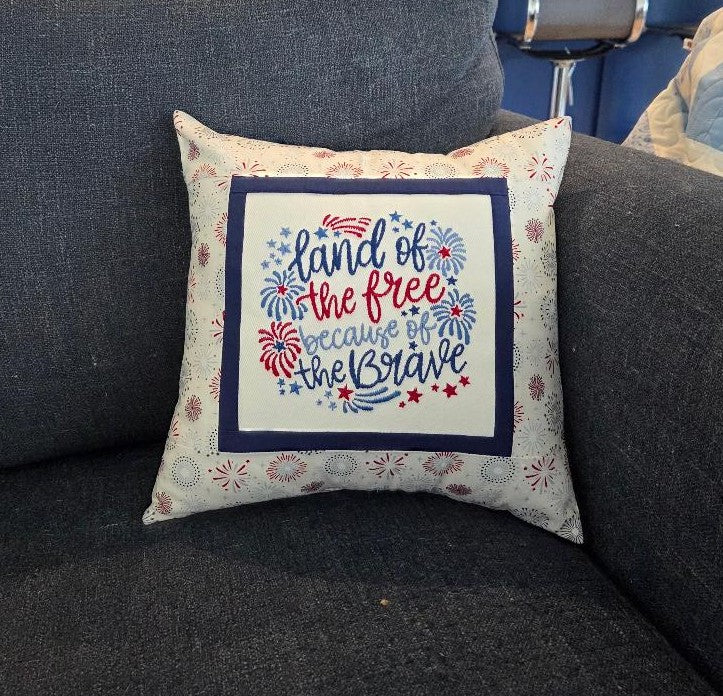 Decorative pillow with 'land of the free because of the brave' text on a gray couch.